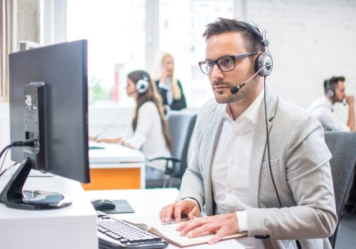 Serious man wearing formal clothes and headset looking at computer screen in bright office